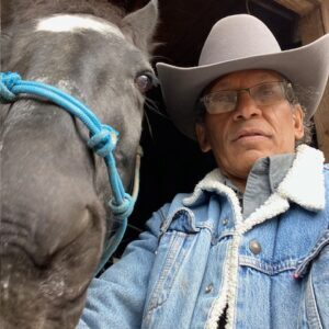 A person wearing a denim jacket, glasses, and a cowboy hat stands next to a horse with a blue halter in front of a wooden structure. The person and the horse are looking directly at the camera.