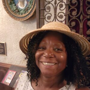 A woman in a straw hat smiles at the camera. She stands indoors against a textured wall with ornate metalwork and a decorative plaque featuring a relief portrait.