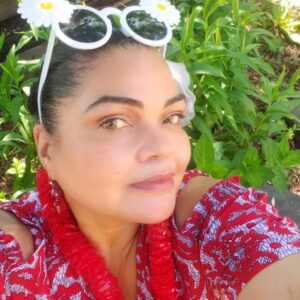 A woman wearing a red patterned outfit and a red lei poses outdoors. She has sunglasses with white daisy decorations. The background features green foliage and a wooden board.