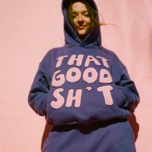 A person wearing a blue hoodie with large white letters spelling THAT GOOD SH*T stands against a pink background. The hoodie is pulled up over their head, and hands are in the pockets. Wind gently blows some hair across their face.