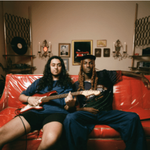 Two people sitting on a red couch in a room with retro decor. One is holding an electric guitar, while the other has an arm on the couch. Walls have framed pictures, candle holders, and a record. Ambient lighting creates a cozy vibe.