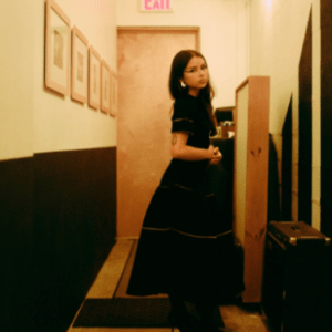 A woman in a black dress stands in a narrow hallway with framed pictures on the wall. She is looking at the camera, with a door and exit sign behind her. The lighting is warm.