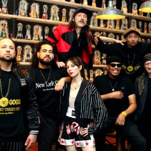 A diverse group of eight individuals poses together in a lively setting. They are dressed in stylish, urban attire, featuring a mix of bold colors and patterns, including pinstripes and graphic designs. The background showcases a wall adorned with colorful figurines, adding a vibrant touch to the scene. The group exudes a confident and energetic vibe, with some members standing and others seated, all engaging with the camera.