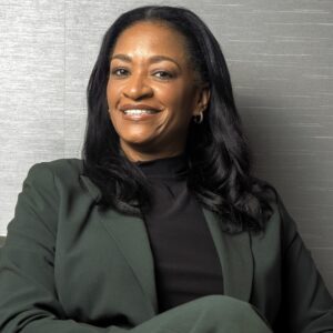A woman is seated in a chair, wearing a dark green suit over a black top. She has long, straight black hair and is smiling confidently. The background features a textured gray wall, adding a professional ambiance to the setting. Her posture is relaxed, with one leg crossed over the other.