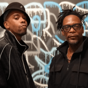 Two people stand in front of a wall with abstract spray-painted graffiti. Both wear black jackets; one has a black hat and the other has black glasses. They appear confident and serious.