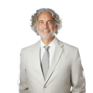 A man with curly gray hair and a beard is wearing a light-colored suit and a tie with a subtle pattern. He is smiling and standing against a plain background. The overall appearance is professional and approachable.