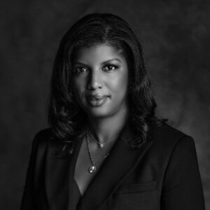 A woman is depicted in a black and white portrait, wearing a tailored black blazer. Her hair is styled in loose waves, framing her face. She has a confident expression, with subtle makeup highlighting her features. Around her neck, she wears two delicate necklaces, one with a star pendant and the other with a circular charm. The background is softly blurred, emphasizing her presence in the foreground.
