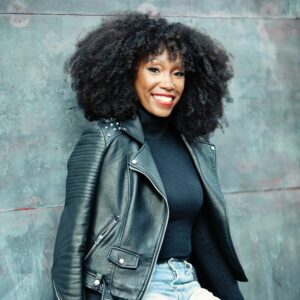 Smiling person with curly hair wearing a black leather jacket, black top, and distressed jeans sits against a textured gray background.