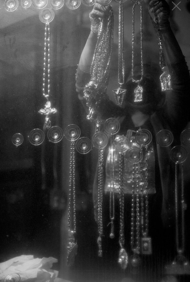 A person holds up necklaces in front of a display, with various pendants and chains visible. The scene is captured in black and white, creating a reflective effect on the glass.