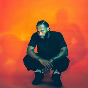 Man squatting in front of an orange background, wearing a black t-shirt, pants, and shoes. His hands are clasped, and his head is slightly tilted, creating a shadow on the backdrop.