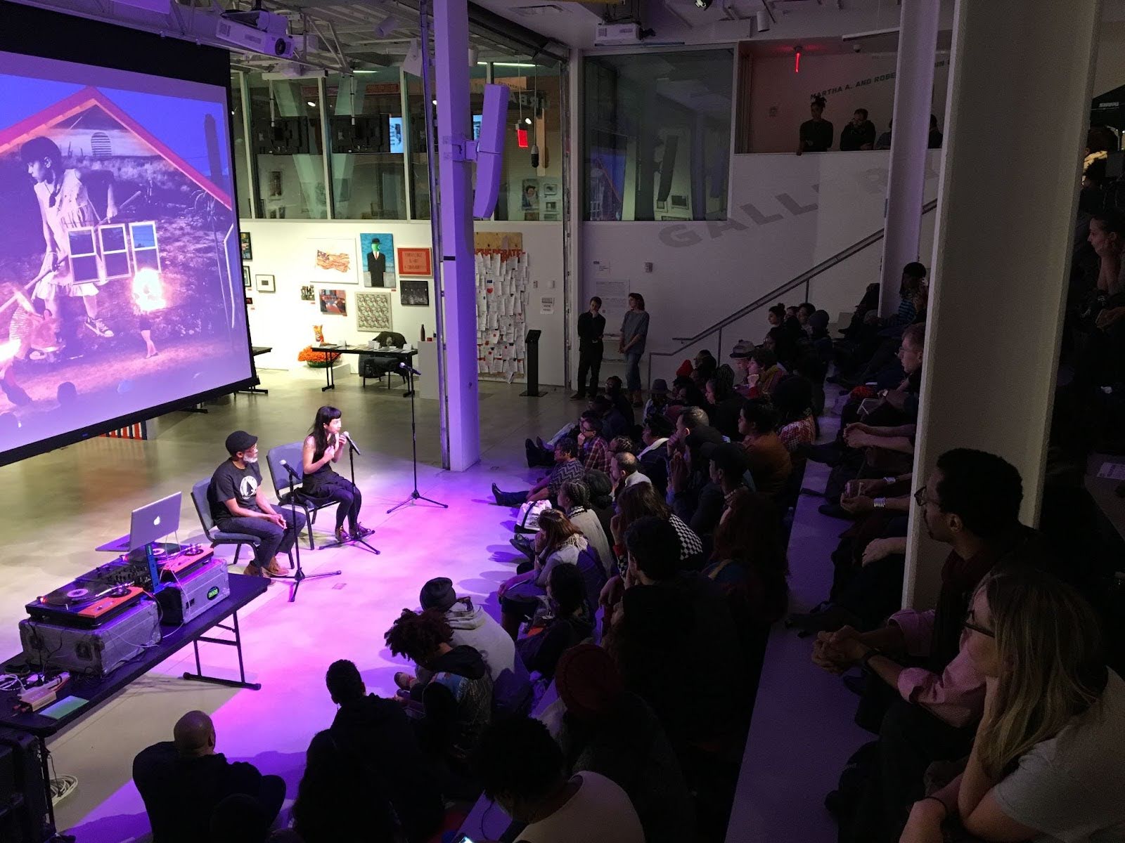 A large group of people sit on steps in a modern indoor space, watching two individuals speak on a stage with microphones. A slide with colorful images is projected on a screen behind them. A table with equipment is beside the stage.