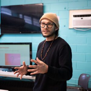 A person with long hair and a beard is standing in front of a turquoise wall, gesturing with their hands as they speak. They are wearing a black sweater and a beige beanie, along with a beaded necklace. In the background, there are two air conditioning units and a television displaying a presentation slide with a question mark on it, labeled "Business Discussion." A table is visible in the foreground, and a chair is positioned nearby.
