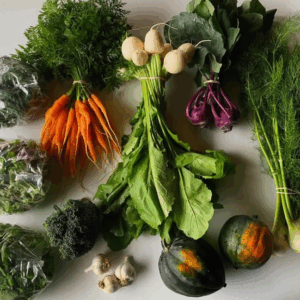 A variety of fresh vegetables is displayed on a light surface. The arrangement includes vibrant orange carrots with green tops, white turnips, purple kohlrabi, and leafy greens. There are also wrapped bundles of mixed salad greens, broccoli, and a bunch of dill. Two acorn squashes, one dark green and one with orange markings, are positioned at the bottom, alongside a few garlic cloves. The overall composition showcases a colorful assortment of produce.